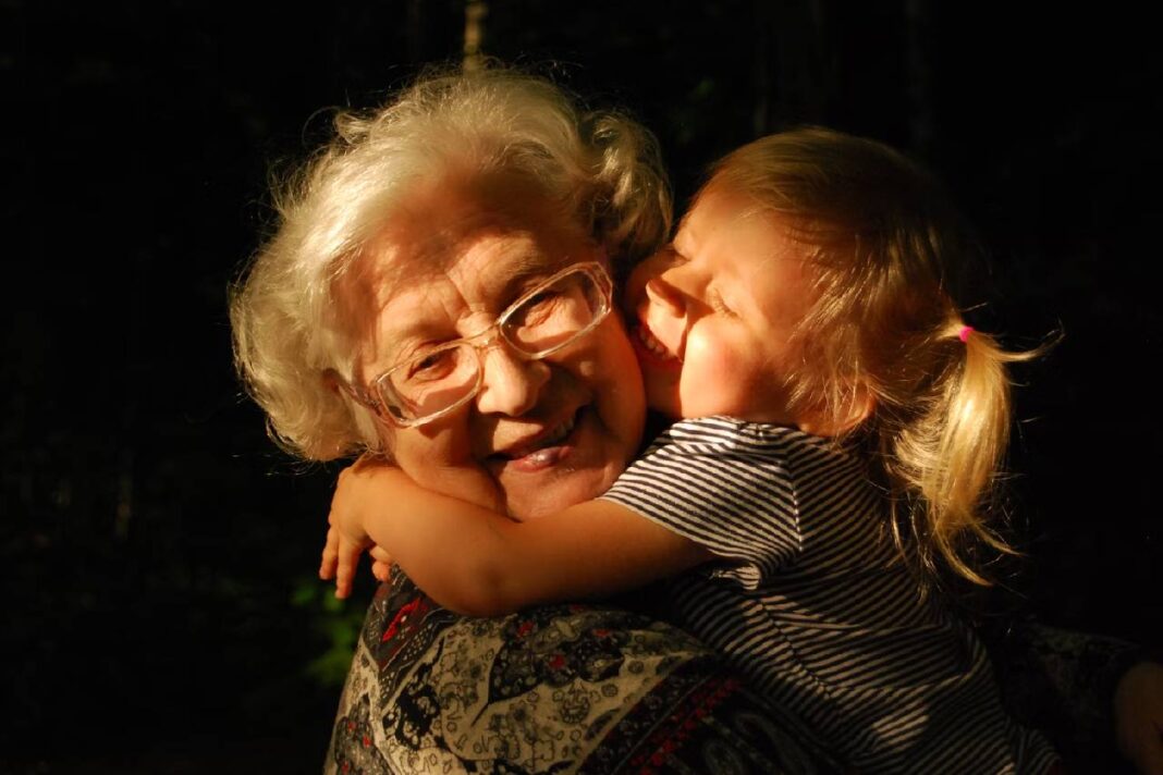 An elderly woman with white hair and glasses smiles warmly as a young blonde girl hugs her tightly, both bathed in golden sunlight.